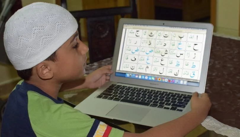 Boy in a prayer cap studying an Arabic letters chart on a laptop during a Noorani Qaida course online