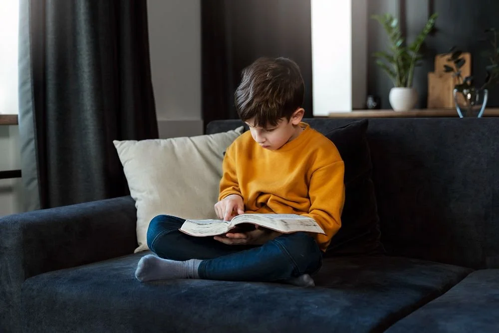 Boy sitting on a sofa reading an open book, illustrating flexible online Quran courses at home